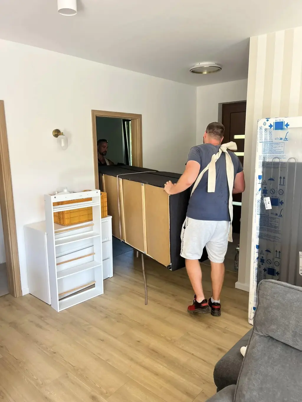Két költöztető munkatárs egy nagy szekrényt visz be egy világos, üres lakásba